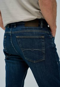Acercamiento de una persona que lleva puestas unas jeans de mezclilla azul oscuro con costuras visibles y una etiqueta de marca marrón en la cinturilla, con las manos en los bolsillos.