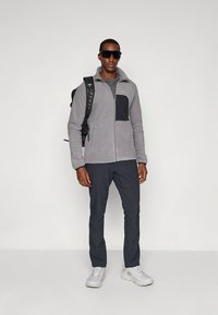 Casaco cinzento de tecido polar com um bolso preto no peito, combinado com calças escuras e ténis brancos. O conjunto é completado com óculos de sol e uma mochila.