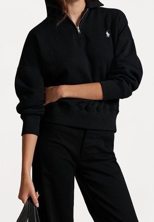 Persoon draagt een zwart sweatvest met kwart rits en wit polo-logo, zwarte broek, en houdt een zwart gestructureerd handtasje naast zich.