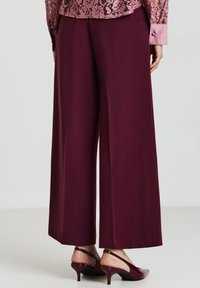 Femme portant un pantalon bordeaux à jambes larges avec des escarpins à bride arrière en cuir verni assortis et un haut en dentelle rose à manches longues, debout sur un sol blanc.