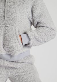 Sudadera de forro polar gris difuso con un bolsillo central, puños y cinturilla acanalados en gris. Cuenta con una capucha con cordón y una superficie texturizada.