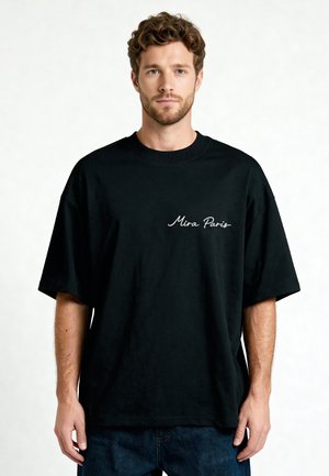 Man met kort krullend haar en baard die een zwarte oversized T-shirt draagt met "Mira Paris" in het wit op de borst gedrukt.