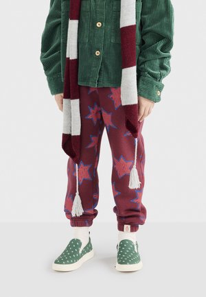 Kind in een groene corduroyjas, gestreepte bordeauxrode en witte sjaal, rode broek met sterrenpatroon, witte sokken en groene instappers.