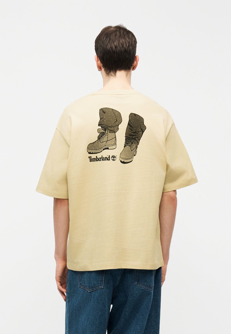 Άτομο που φοράει μπεζ φαρδιά μπλούζα με γραφικό απεικονιζόμενο με ανασηκωμένες μπότες και το λογότυπο "Timberland" στην πλάτη, σε συνδυασμό με μπλε τζιν.