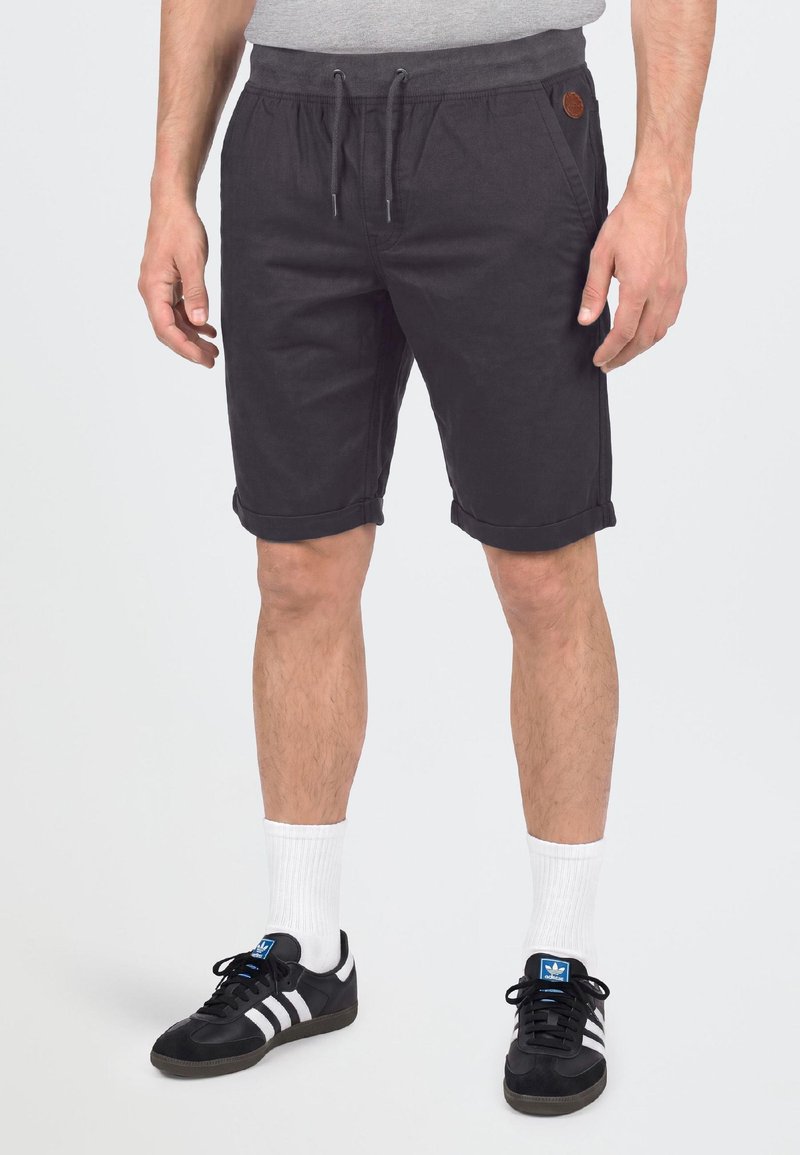 Homme portant un short noir à cordon, des chaussettes blanches montantes et des baskets Adidas noires avec des bandes blanches, debout devant un fond uni.