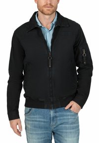 Schwarze Bomberjacke mit einem Reißverschluss vorne, gerippten Bündchen und Saum; hat einen strukturierten Stoff und seitliche Reißverschlusstaschen. Getragen über einem hellblauen Hemd.