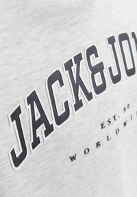 Szara tkanina z dużym czarno-białym obrysem tekstu "JACK & JONES" i mniejszym tekstem poniżej "EST. 90 WORLDWIDE".