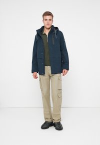 Νεαρός άντρας στέκεται, φορώντας ναυτικό μπλε φούτερ με κουκούλα, ελαιόχρωμο μπουφάν με φερμουάρ, μπεζ cargo παντελόνι και μαύρα αθλητικά παπούτσια, με φόντο λευκό. 