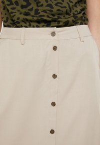 Beiger Rock aus leichtem Stoff, mit einer Knopfleiste vorne und braunen Knöpfen sowie einer glatten Textur. Schlichtes Design mit Gürtelschlaufen.