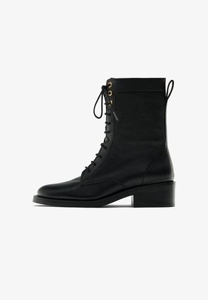 Bottine en cuir noir avec lacets sur le devant, œillets dorés, bout arrondi, talon bloc de 4 cm, et languette arrière, sur fond blanc.