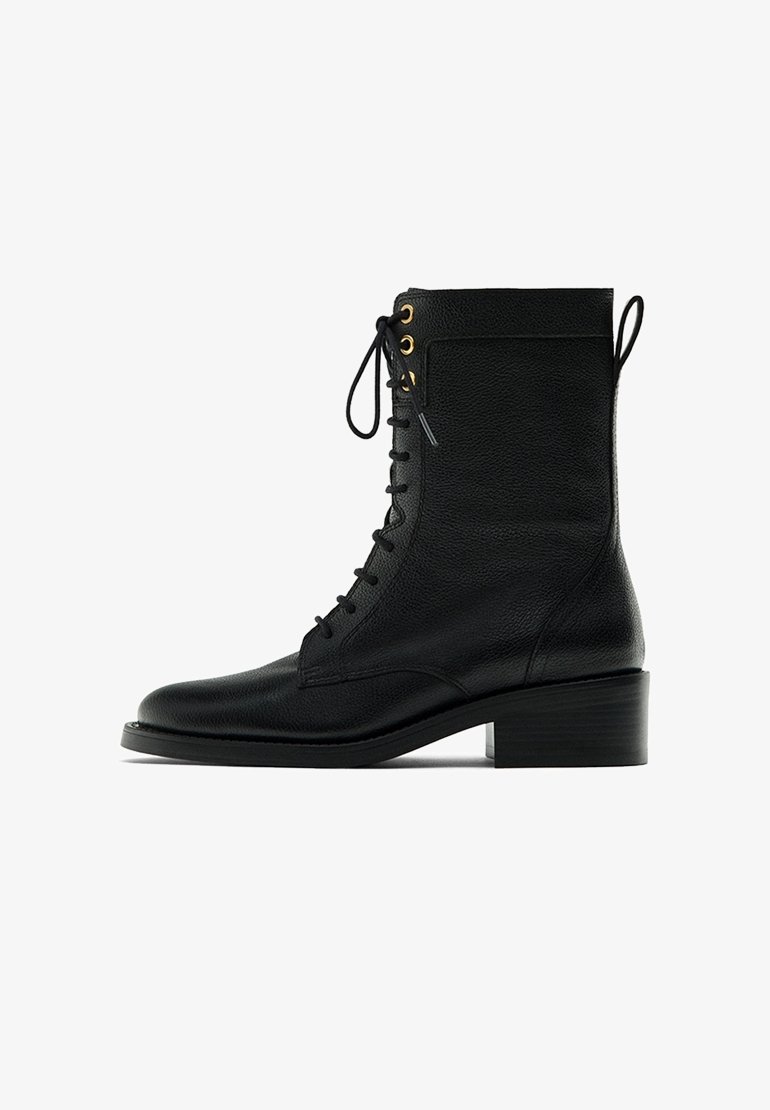 Bottine en cuir noir avec lacets sur le devant, œillets dorés, bout arrondi, talon bloc de 4 cm, et languette arrière, sur fond blanc.