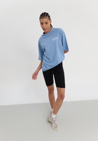 Blå oversized t-skjorte med «ARCHIVE» trykk, paret med svarte sykkelshorts og lyse joggesko. Glatt stoff med avslappet passform.