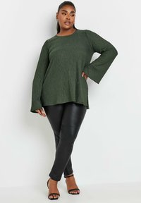 Top verde scuro con texture e maniche lunghe, abbinato a pantaloni aderenti simili al cuoio neri e tacchi a spillo neri.