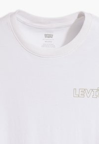 Camiseta de algodón blanca con cuello redondo, que presenta un logo en relieve "LEVI'S" en gris claro en el lado izquierdo del pecho. Textura suave, corte relajado.