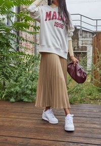 Ljusgrå sweatshirt med "MALI 1996" i burgundy, beige plisserad kjol, vita sneakers och en burgundy clutch mot en grön bakgrund.
