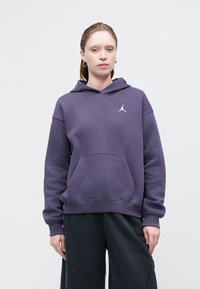 Sudadera con capucha morada, con un bolsillo tipo canguro y un pequeño logo blanco en el pecho, combinada con pantalones negros sueltos. Tejido suave y texturizado.