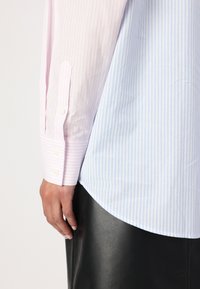 Striped shirt with pink and light blue sections, featuring buttoned cuffs and a curved hemline, worn over a black textured skirt.
