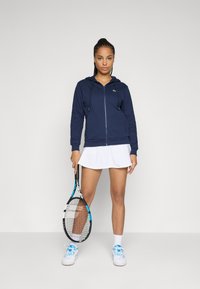 Marineblå hettegenser med glidelås, sammen med et hvitt treningsskjørt. Modellen holder en tenniskølle, iført hvite joggesko med blå detaljer.