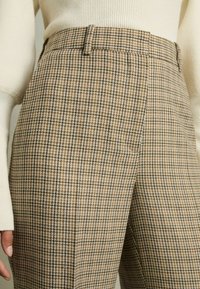 Bruna rutiga byxor med hög midja, med en texturerad tyg, bältesöglor och sidofickor. Mönstret innehåller jordnära toner.