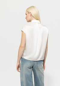 Cremefarbige Seidenbluse mit kurzen Cap-Ärmeln, einem Kragen und einer Mittelnaht auf der Rückseite, kombiniert mit hellblauen weit geschnittenen Jeans.