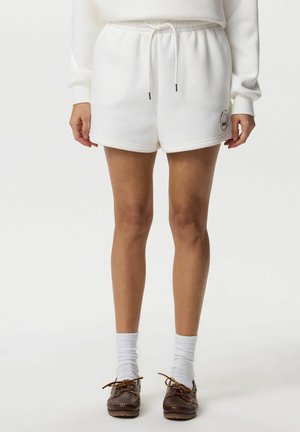 Osoba nosząca białe szorty ze ściągaczem, białe skarpetki i brązowe, sznurowane mokasyny, stojąca na jednolitym tle.