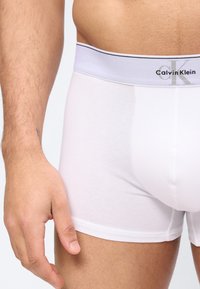 Άνδρας που φοράει λευκά μποξέρ Calvin Klein, ορατός από τη μέση μέχρι το μέσο του μηρού, με το αριστερό χέρι χαλαρό δίπλα στο σώμα.