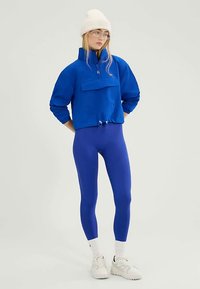 Blauwe pullover met halve rits, voorzak en trekkoordzoom, gecombineerd met geribbelde blauwe legging en witte sportschoenen. Gebreide witte beanie.