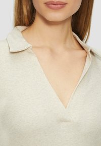 Beżowy, prążkowany top z kołnierzykiem i dekoltem w kształcie litery V. Charakteryzuje się miękką teksturą i detalami przeszycia wzdłuż kołnierza.