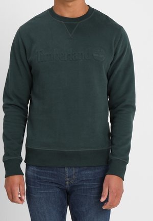 Mężczyzna w ciemnozielonej bluzie Timberland i niebieskich dżinsach, stojący na jednolitym jasnym tle.