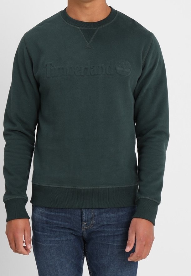 Homme portant un sweat Timberland vert foncé et un jean bleu, debout devant un fond clair uni.