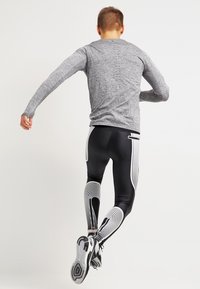 Sportoutfit voor mannen met een grijze lange mouw top en zwarte leggings met witte golvende strepen, ontworpen voor prestaties en flexibiliteit.