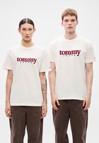 Krótki rękaw, kremowe koszulki z logo "tommy JEANS" w kolorze czerwonym. Model po lewej ma tatuaże; obaj mają na sobie ciemnobrązowe spodnie.