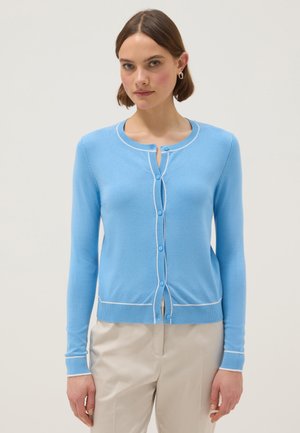 Mujer vistiendo un cárdigan azul claro con ribetes blancos y pantalones beige, de pie contra un fondo claro y liso.