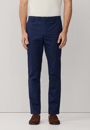 Homme portant un pantalon bleu marine slim, un t-shirt blanc et des mocassins en daim marron, debout devant un fond clair uni.