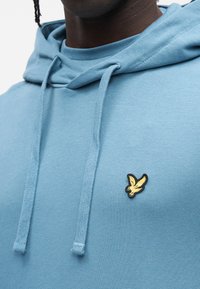 Sudadera azul con capucha hecha de tela suave con un bolsillo tipo canguro, presenta un logo bordado en negro y dorado en el pecho y cordones en la capucha.