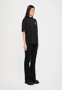 T-shirt nera in cotone con una vestibilità rilassata e maniche corte, caratterizzata da un piccolo design grafico sul petto. Abbinata a pantaloni neri a zampa.