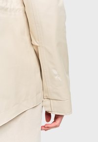 Manche de veste beige avec un petit logo éclair réfléchissant près du poignet, portée par-dessus un pantalon de couleur similaire.