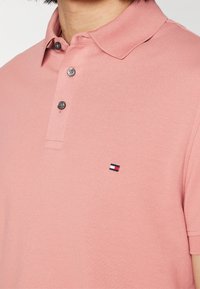 Ljusrosa pikétröja av strukturerat tyg, med klassisk krage, tre knappar och en liten broderad logga på bröstet.