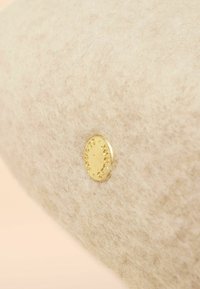 Bézs színű filcanyag puha tapintással, egyik oldalán kör alakú, aranyszínű fém díszítéssel, amely dombornyomott szöveget tartalmaz.