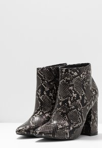 Un paio di stivali alla caviglia in pelle di serpente nera e grigia con punte affusolate e tacchi a blocco su superficie bianca.