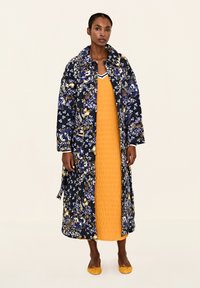 Langer Mantel mit floralem Muster in Schwarz, Blau und Gelb über einem strukturierten gelben Kleid. Helle gelbe Ballerinas runden das Outfit ab.
