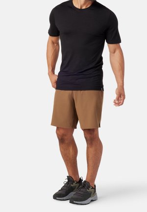 Homme portant un t-shirt noir à manches courtes, un short marron au genou et des chaussures de sport noires, debout devant un fond blanc.