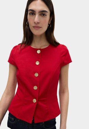 Donna che indossa un fitted top rosso a maniche corte con cinque grandi bottoni texturizzati dorati sulla parte anteriore e orecchini a cerchio dorati.