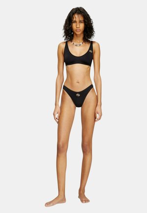 Donna in piedi a piedi nudi che indossa un costume da bagno nero a due pezzi con spalline sottili e slip a vita alta su uno sfondo chiaro e uniforme.