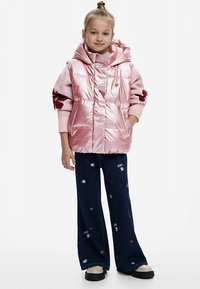 Lyserød polstret vest med hætte, kombineret med en lyserød sweater og mørkeblå vidde-bukser med blomsterbroderi. Sorte plateau-sko.