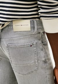 Grijze denim jeans hebben een leren patch met het "TOMMY HILFIGER" logo, achterzakken met kleine embleemaccenten en een slanke, aansluitende pasvorm.