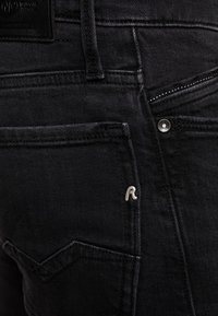 Schwarze Jeans aus Denim mit sichtbaren Nähten und einem kleinen silbernen "R"-Logo. Verfügt über eine Vorderseitentasche und einen Reißverschluss an der rechten Seite.