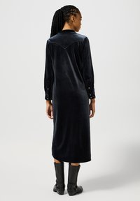 Lungo abito di velluto nero con colletto, polsini con bottoni e un dettaglio a yoke a forma di V sul retro. Indossato con stivali neri al ginocchio.