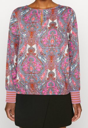 Kobieta ubrana w bluzkę z długim rękawem w różowe, pomarańczowe i niebieskie wzory paisley oraz w paski na mankietach, w połączeniu z czarną spódnicą.