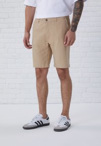 Beige cotton shorts with a button closure, side pockets, and a straight fit, paired with white and gray athletic shoes featuring black stripes.